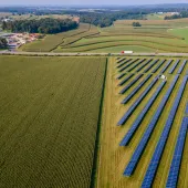 Aerial view of a rural landscape with fields of crops and rows of solar panels. A small settlement is visible in the distance, surrounded by rolling hills.