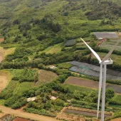 Aerial view of two wind turbines situated on a lush green landscape with surrounding farmland. The turbines are positioned on a dirt path among various agricultural plots, with dense forested hills in the background.