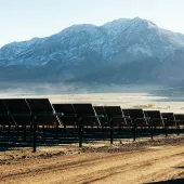 Rows of solar panels on a dirt field with a backdrop of snow-capped mountains under a clear sky.