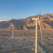 Aerial view of wind turbines in a desert landscape with mountains in the background under a clear blue sky.