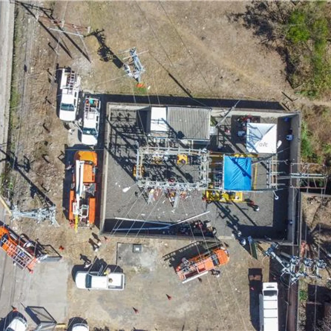 Aerial view of a power substation with multiple vehicles and equipment around it. The substation is surrounded by a fence and located near a road. The area is dry with some vegetation nearby.