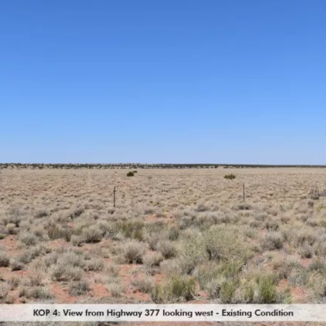 A flat, arid landscape with sparse vegetation and a clear blue sky, viewed from Highway 377 looking west. The ground is covered with dry grass and small shrubs, and a barbed wire fence runs across the scene.