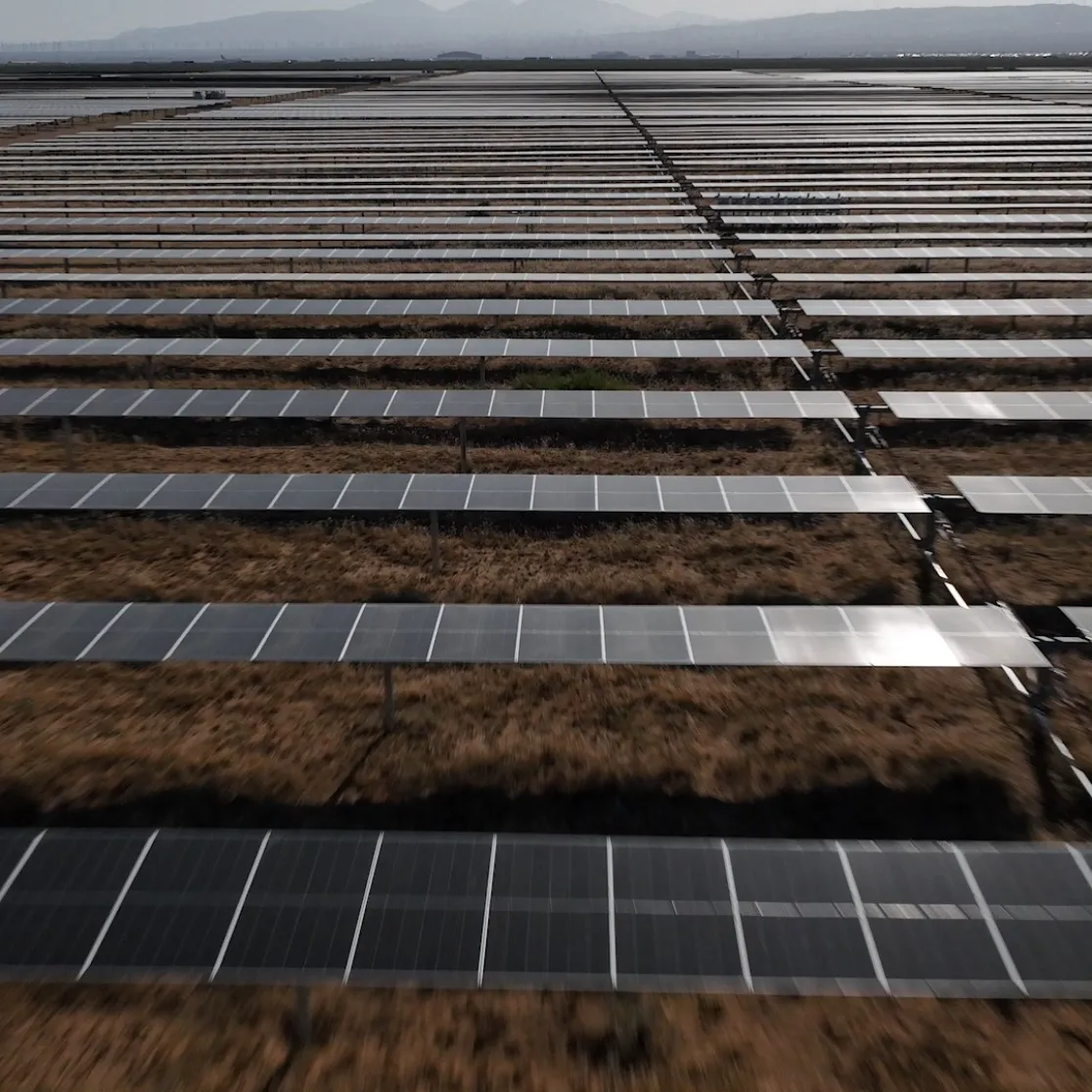 A large field of solar panels arranged in parallel rows, stretching into the distance. The panels are set on a dry, grassy terrain, with mountains visible on the horizon under a cloudy sky.