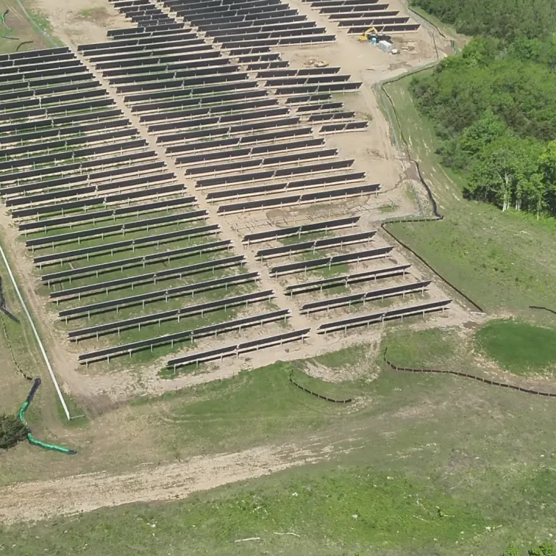 Aerial view of a solar farm with rows of solar panels installed on a cleared piece of land surrounded by dense green forest. The panels are aligned in parallel rows, and there are patches of grass and dirt visible around them.