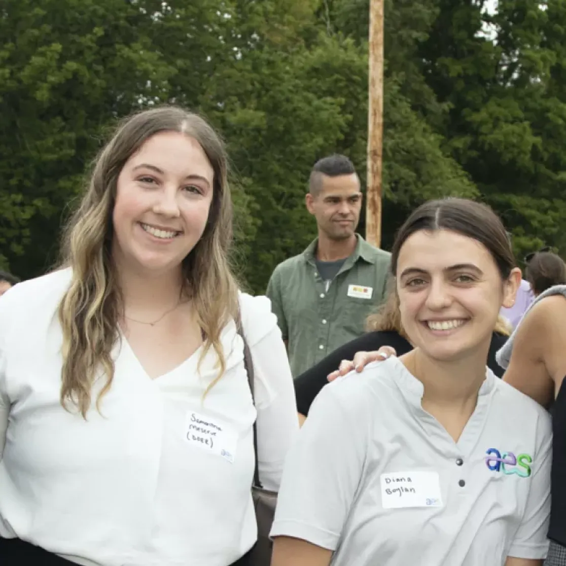 Five people standing outdoors smiling at the camera. They are wearing name tags and casual clothing. Trees and other people are visible in the background.