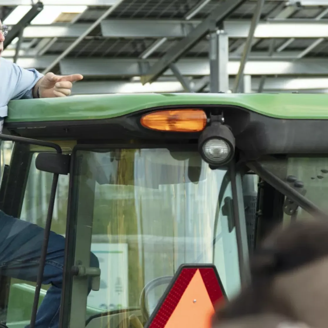 An older man in a blue shirt stands on a green tractor, pointing and speaking to a group of people. The background shows a structure with metal beams and solar panels.