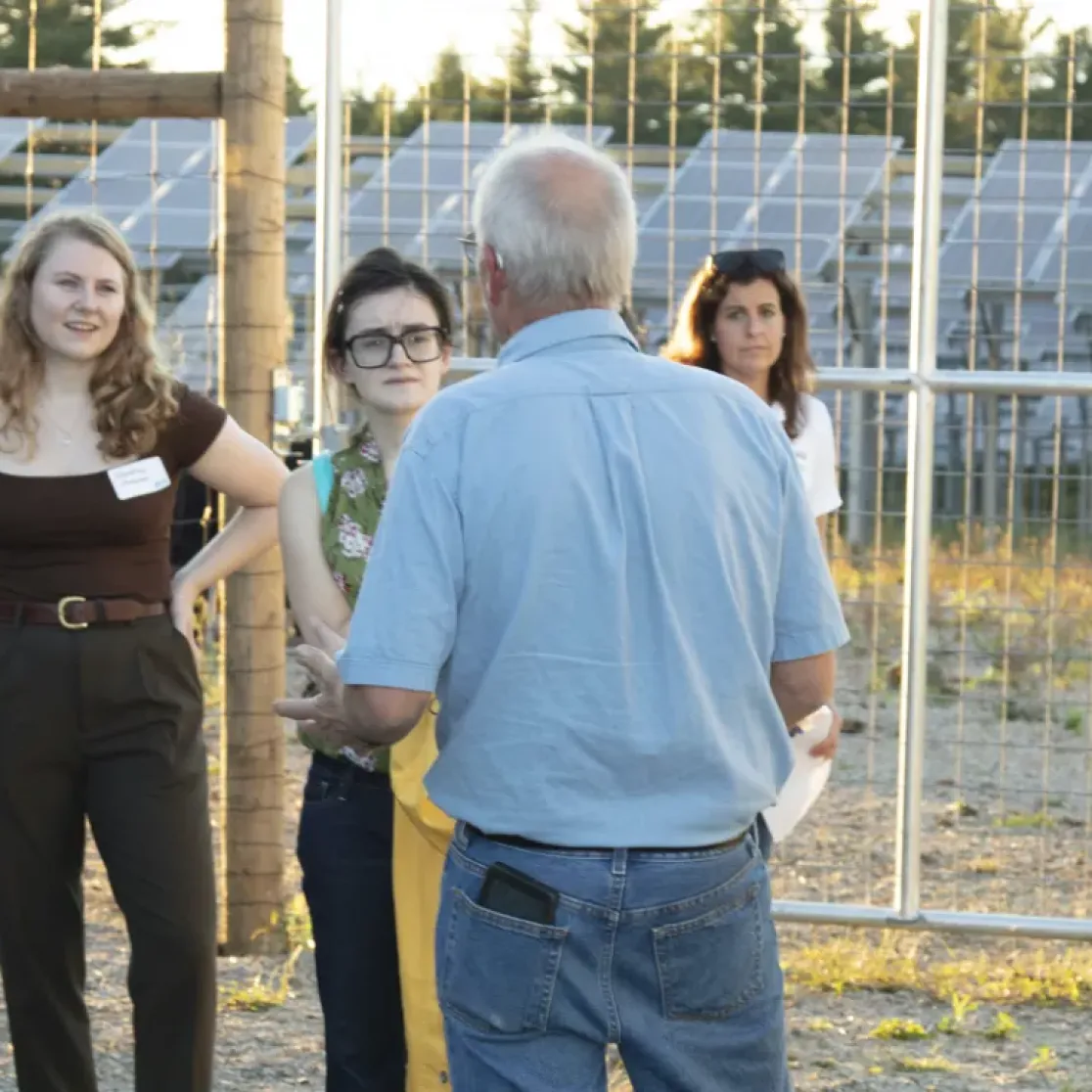 A group of people stands outside near solar panels, engaged in conversation. The setting is sunny, and they are casually dressed. The background features a fence and rows of solar panels.
