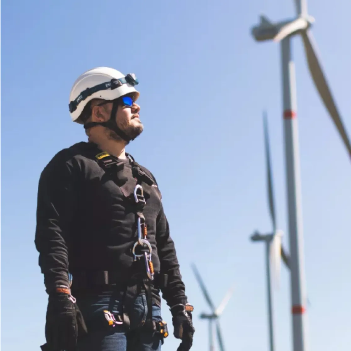 A worker wearing a safety harness, helmet, and sunglasses stands in front of wind turbines under a clear blue sky.