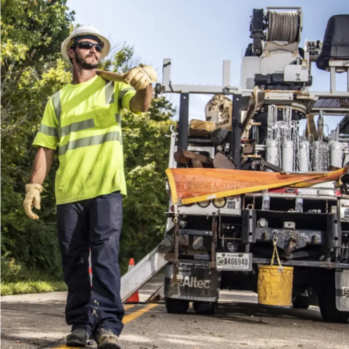 Construction worker in a yellow safety vest and hard hat stands near a utility truck on a road, surrounded by trees. He is wearing gloves and sunglasses, and appears to be giving a signal.