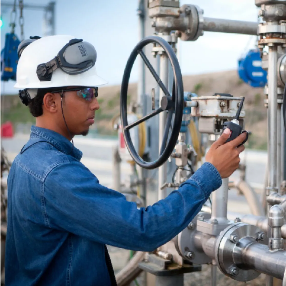 A worker in a hard hat and safety glasses operates a valve at an industrial site, using a handheld device. He is wearing a denim shirt and has ear protection on his helmet.