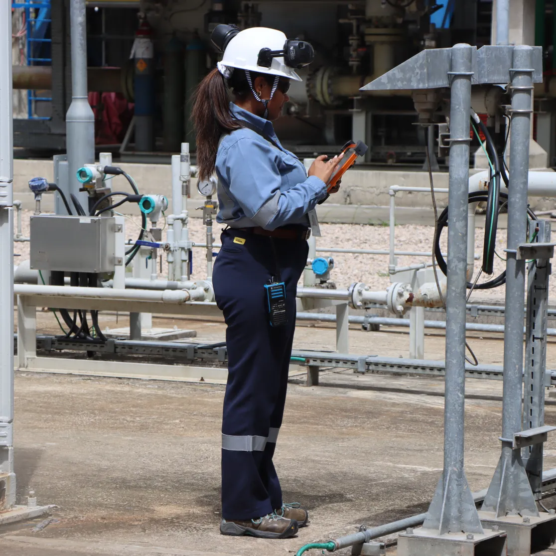 A female industrial technician wearing a hard hat and safety gear, using a handheld digital device at a refinery.