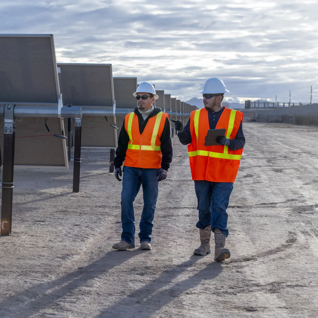 Two solar technicians in hard hats and high-visibility vests walking and talking at a solar farm.