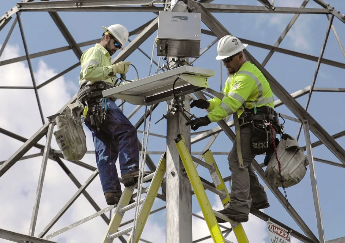 Two workers in safety gear install equipment on a metal tower against a blue sky.
