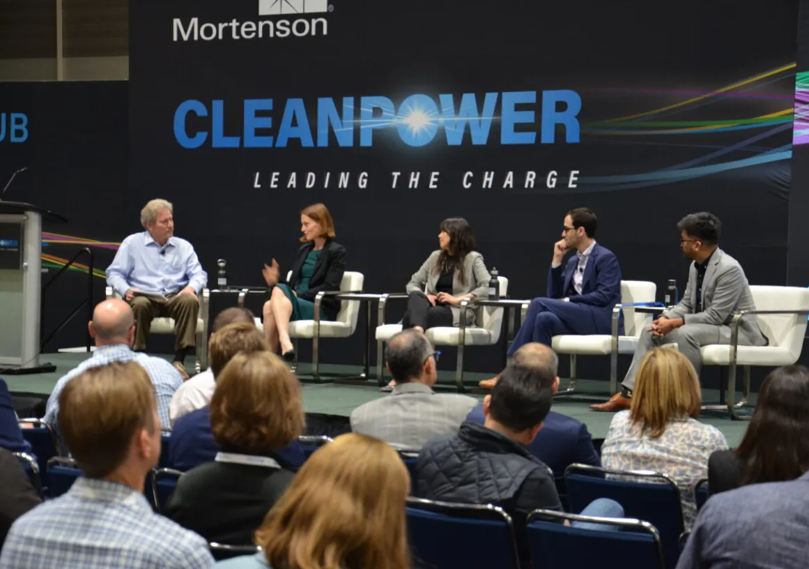 A panel of five people seated on a stage at a CleanPower event, with an audience in the foreground. The backdrop displays the words 'CleanPower Leading the Charge.'