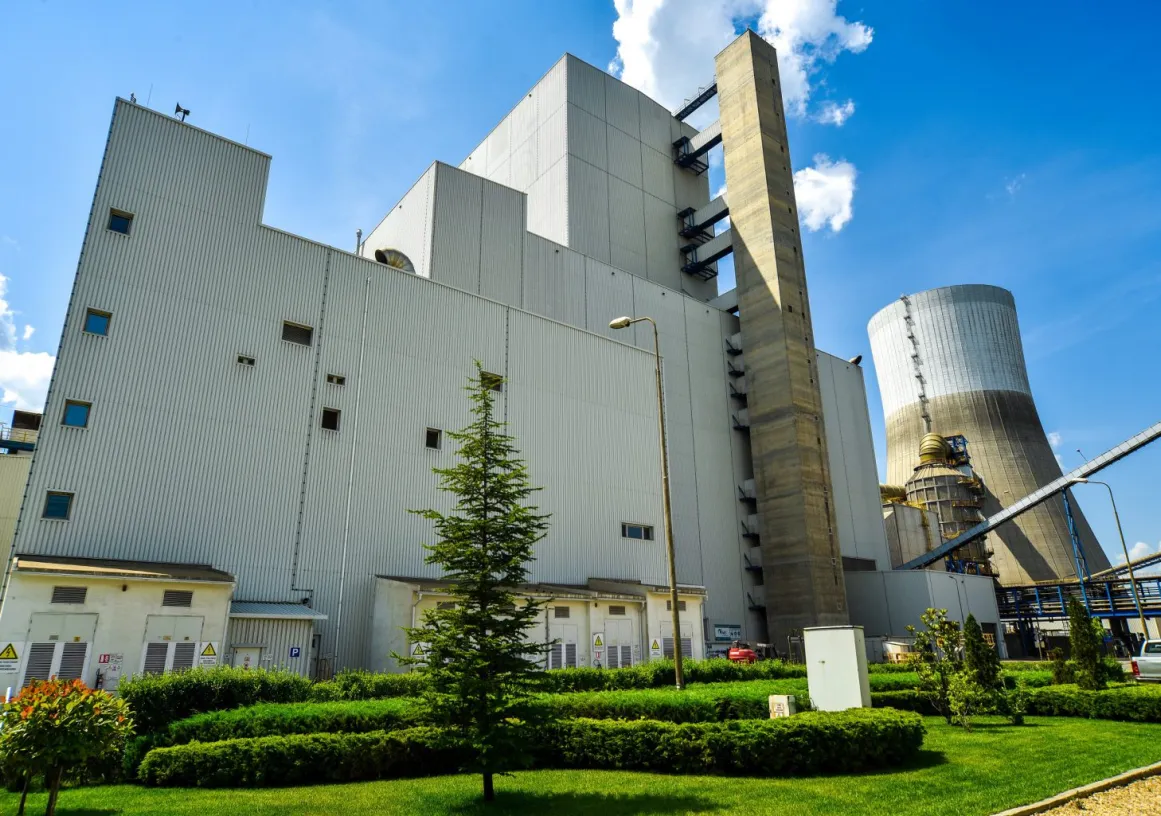 A large industrial building with a corrugated metal exterior, surrounded by green shrubs and trees. A tall smokestack and cooling tower are visible in the background. The sky is clear with a few clouds.