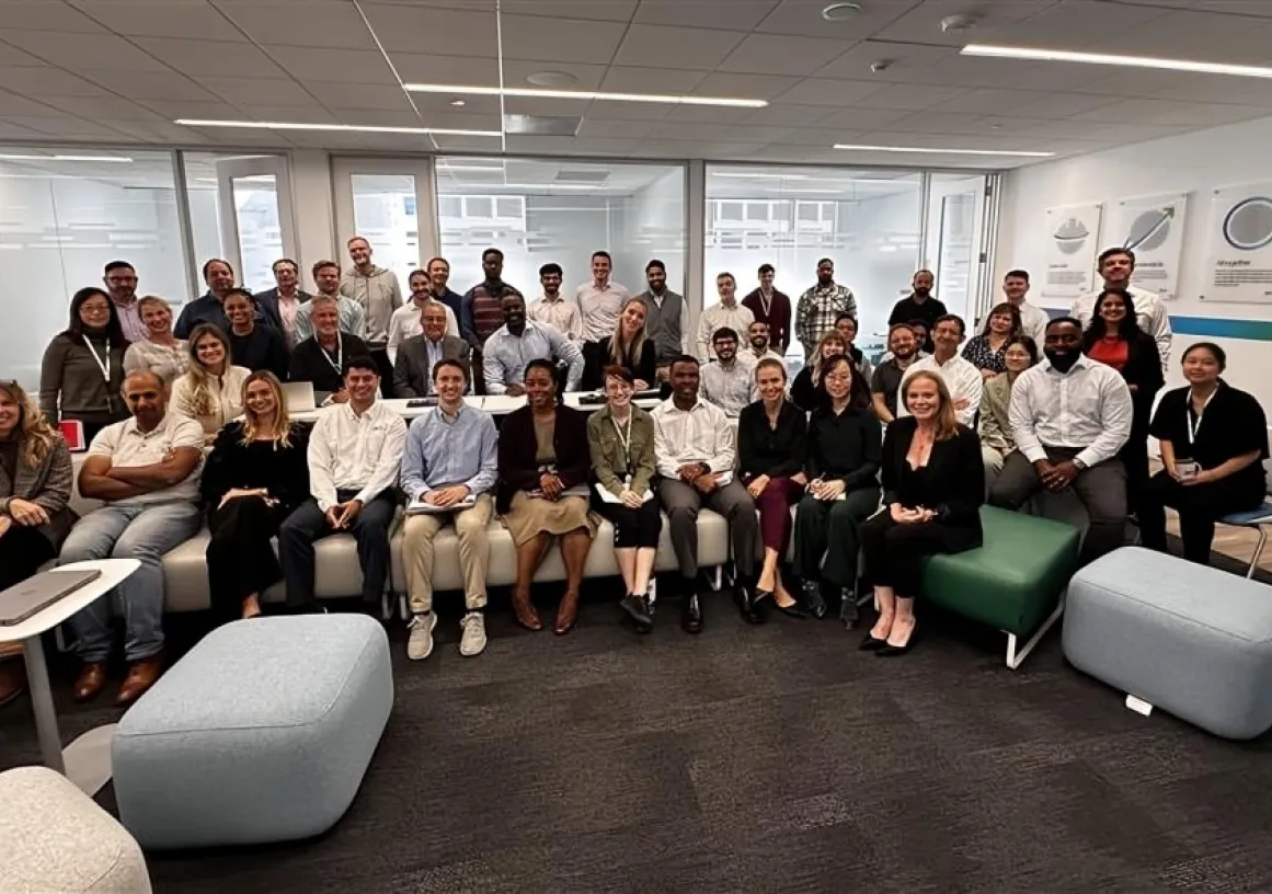 A diverse group of people, approximately 40 individuals, are gathered in an office setting. They are smiling and seated on modern furniture, including sofas and ottomans, with some standing at the back. The room has a contemporary design with glass walls and framed posters on the wall.