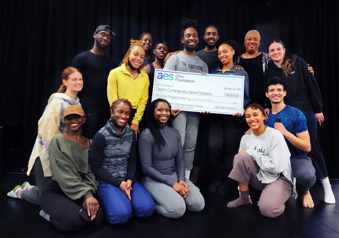 A group of people smiling and posing with a large ceremonial check from the AES Ohio Foundation, made out to the Dayton Contemporary Dance Company for $55,000, dated February 22, 2023. The group is standing and kneeling in front of a dark background.