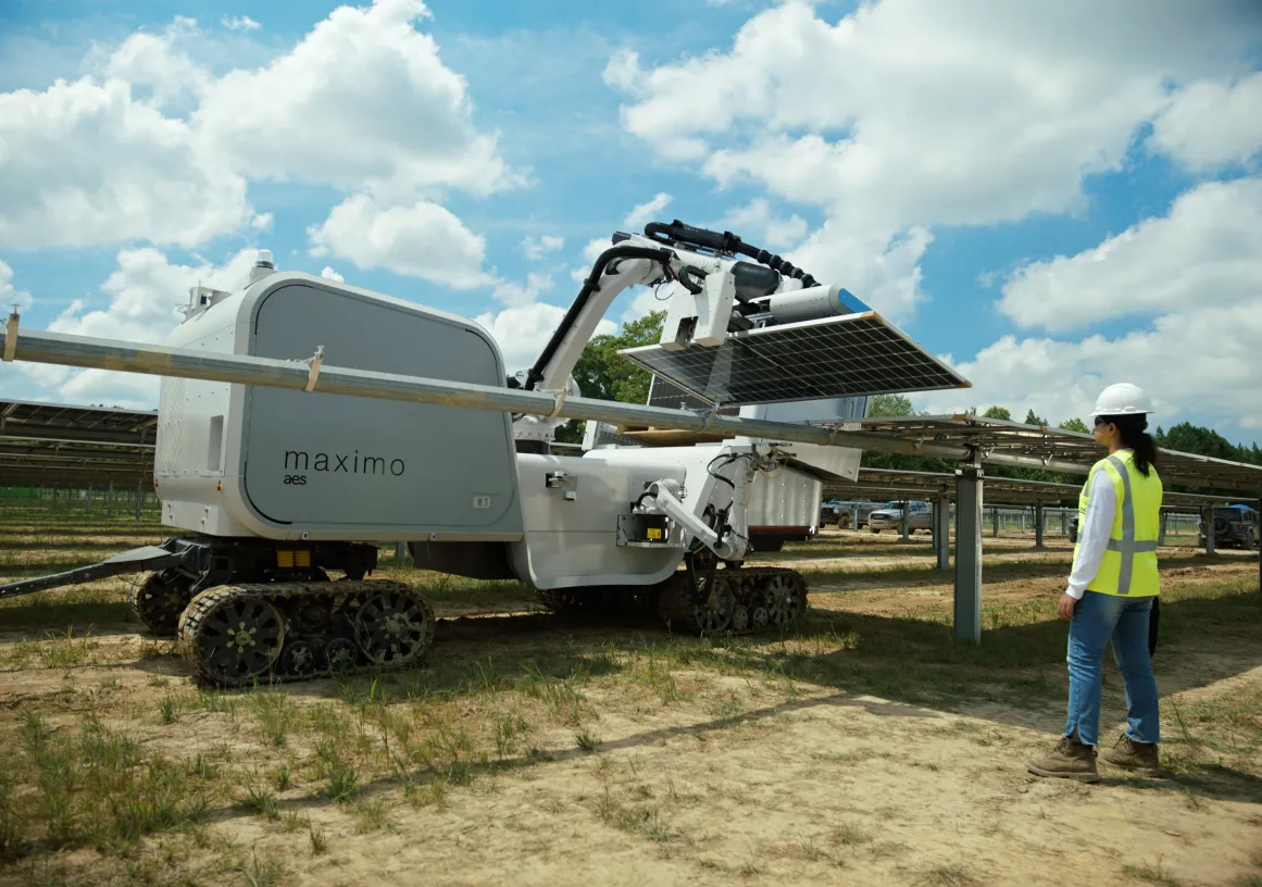 A robotic machine labeled 'maximo aes' is operating in a solar farm, adjusting a solar panel. The machine is on caterpillar tracks, and a person in a safety vest and helmet observes nearby. The background shows rows of solar panels under a partly cloudy sky.