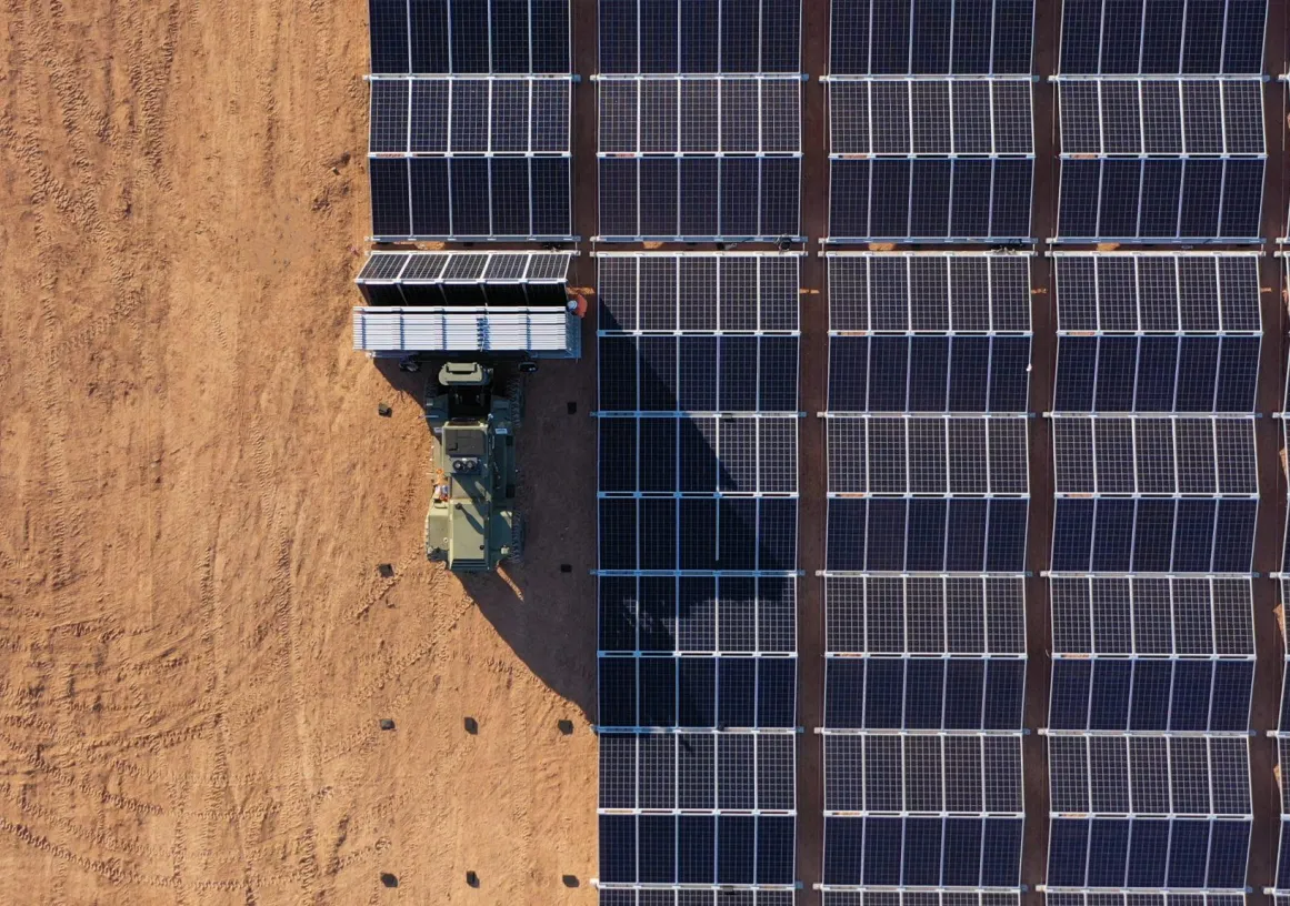 Aerial view of a solar farm with rows of solar panels on the right and a tractor on the left on a dirt field.