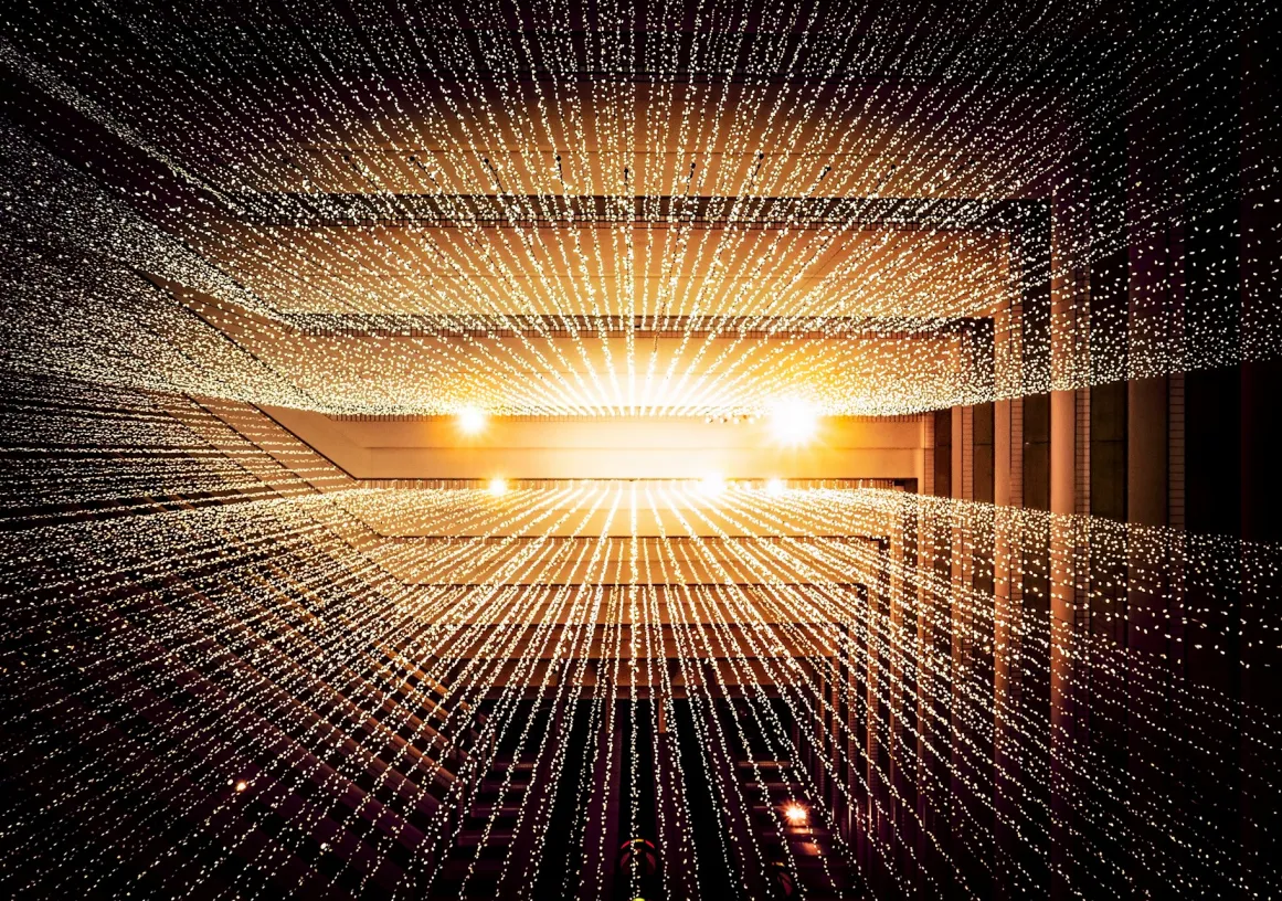 Rows of small white lights suspended in a geometric pattern inside a modern building, creating a radiant, starburst effect with warm lighting in the background.