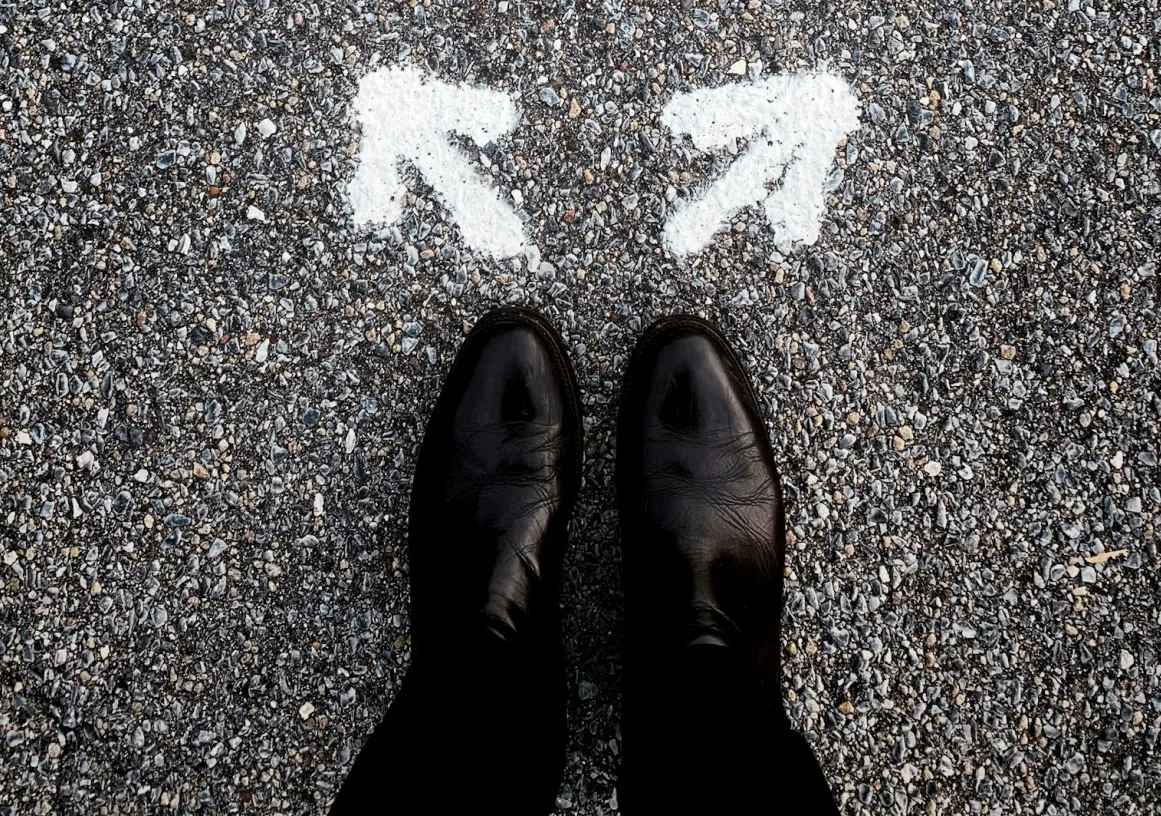 A person wearing black shoes stands on a gravel road in front of two white arrows painted on the ground, pointing in different directions, symbolizing a choice or decision.