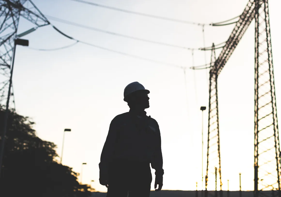 Silhouette of an electrical worker wearing a hard hat standing in front of high voltage transmission towers at sunset.