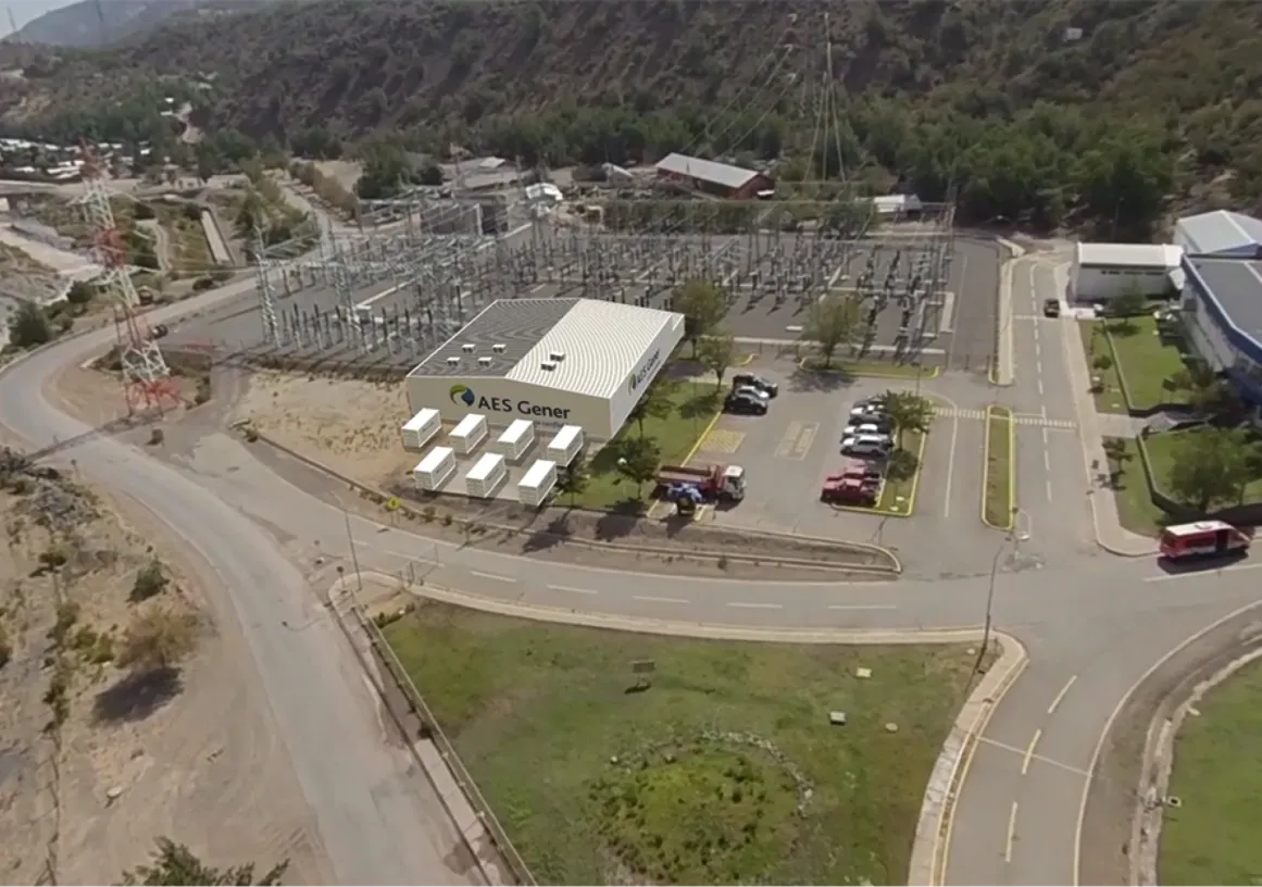 Aerial view of an industrial facility labeled 'AES Gener' surrounded by roads and greenery. The facility includes a main building, parking lot with several cars, and adjacent structures. Nearby, there is a substation with electrical equipment and power lines. The area is set in a mountainous landscape with trees and winding roads.