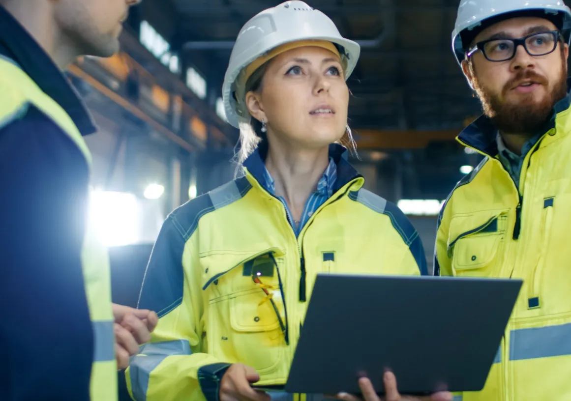Three construction workers wearing safety helmets and high-visibility jackets stand inside a large industrial facility. One holds a laptop, and they appear to be discussing or inspecting something above.