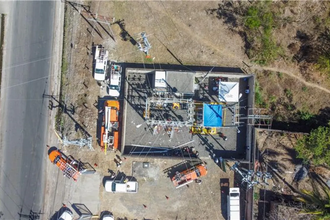 Aerial view of a power substation with multiple vehicles and equipment around it. The substation is surrounded by a fence and located near a road. The area is dry with some vegetation nearby.