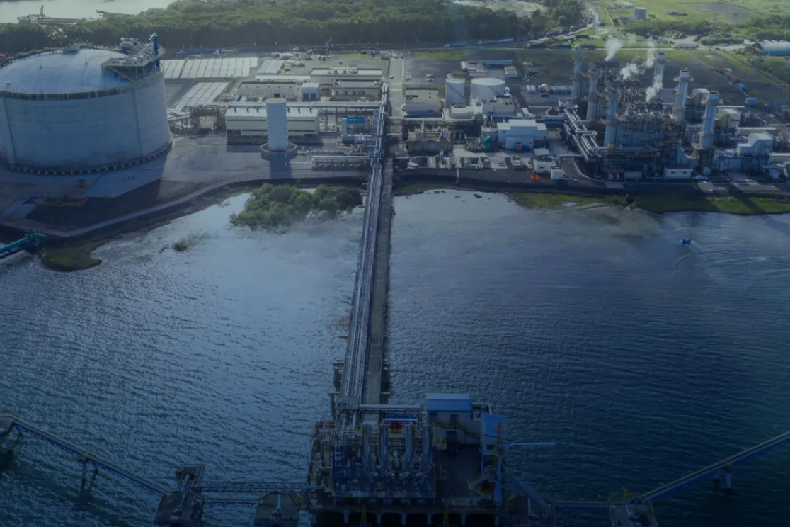 Aerial view of an industrial facility by the water, featuring large storage tanks, pipelines, and buildings. A long pier extends into the water, connecting the facility to the shoreline.