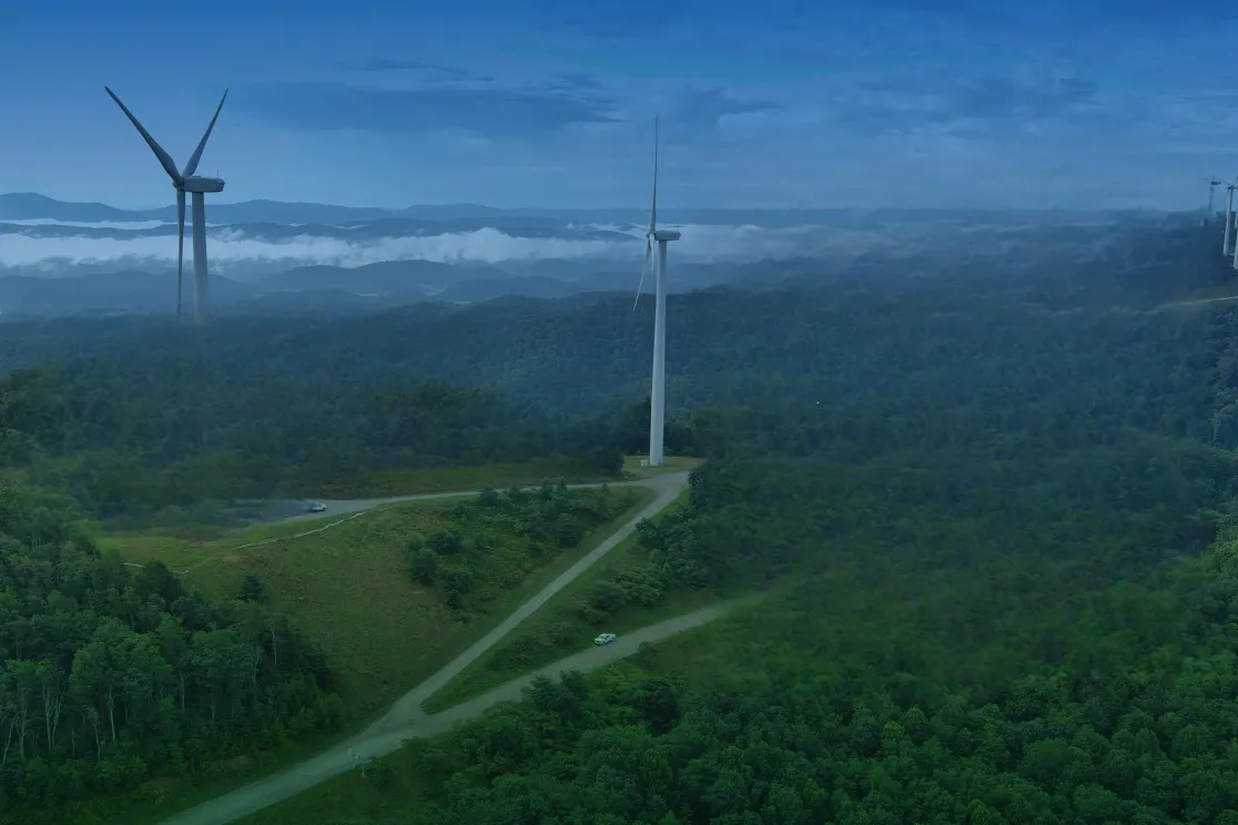 Aerial view of a wind farm with multiple wind turbines on a lush green hilltop, surrounded by dense forests and distant mountain ranges under a clear blue sky.