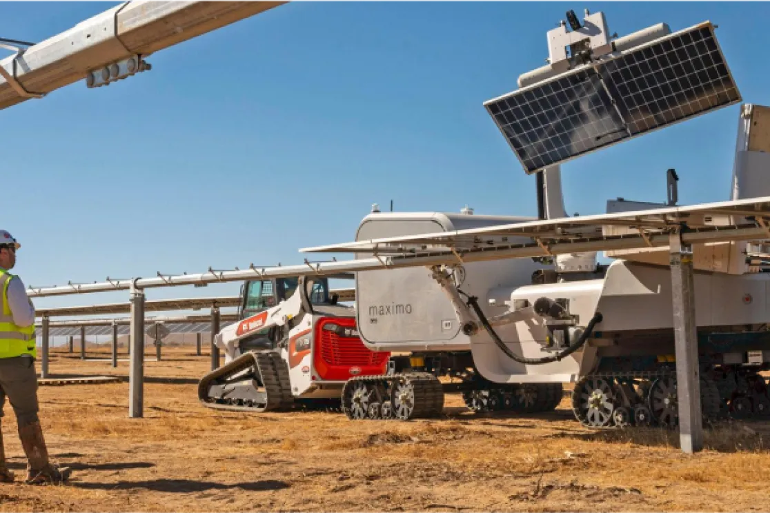 Two workers in safety vests and helmets observe a robotic machine cleaning solar panels in a solar farm under a clear blue sky.