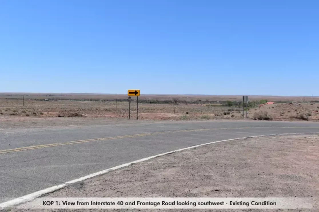 A view from Interstate 40 and Frontage Road looking southwest. The image shows a wide expanse of flat, arid land under a clear blue sky. A road curves in the foreground with a yellow arrow sign indicating a left turn. Sparse vegetation is visible in the distance.