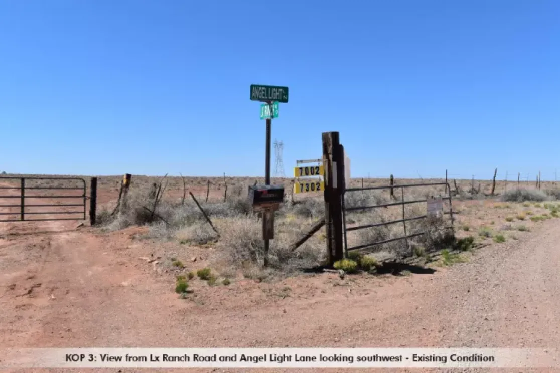 Intersection of Lx Ranch Road and Angel Light Lane in a rural area, looking southwest. Dirt roads intersect with a metal gate on the left and a fence on the right. Road signs display 'Angel Light Ln' and 'Lx Ranch Rd'. Sparse vegetation and clear blue sky are visible.