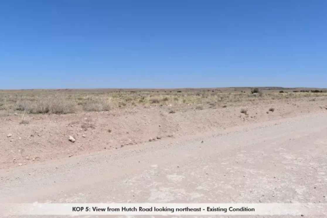 A dry, barren landscape viewed from Hutch Road looking northeast. The foreground shows a dirt road with sparse vegetation on either side. The background features a flat, open expanse under a clear blue sky.
