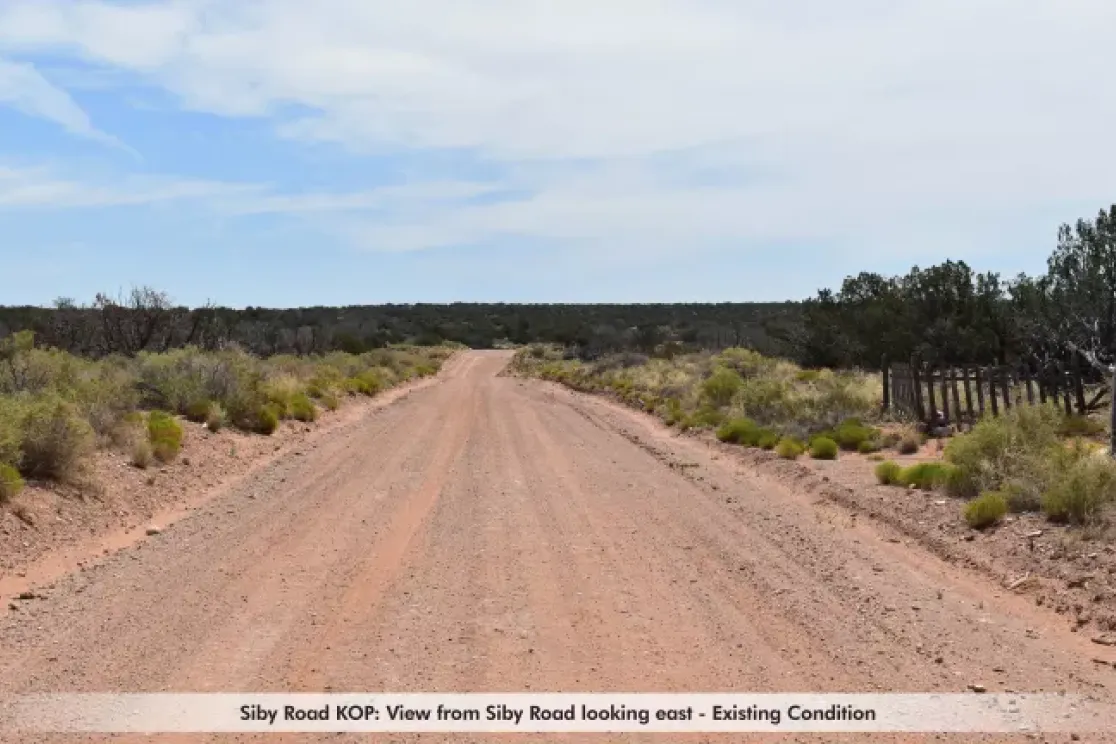 A dirt road stretches into the distance under a blue sky with scattered clouds. The road is flanked by sparse vegetation and low shrubs on both sides. The landscape is flat with a line of trees in the background. A caption at the bottom reads, 'Siby Road KOP: View from Siby Road looking east - Existing Condition.'