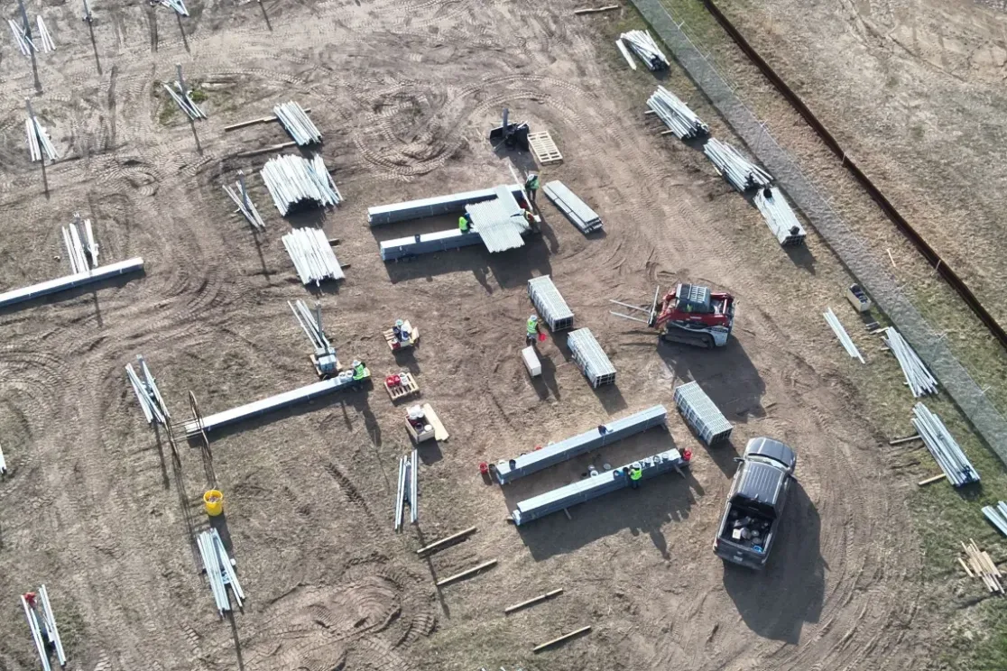 Aerial view of a construction site with scattered metal beams and materials on a dirt field. Workers and machinery are visible, including a red loader and a truck. A small white building with a curved roof is on the right, surrounded by grass.