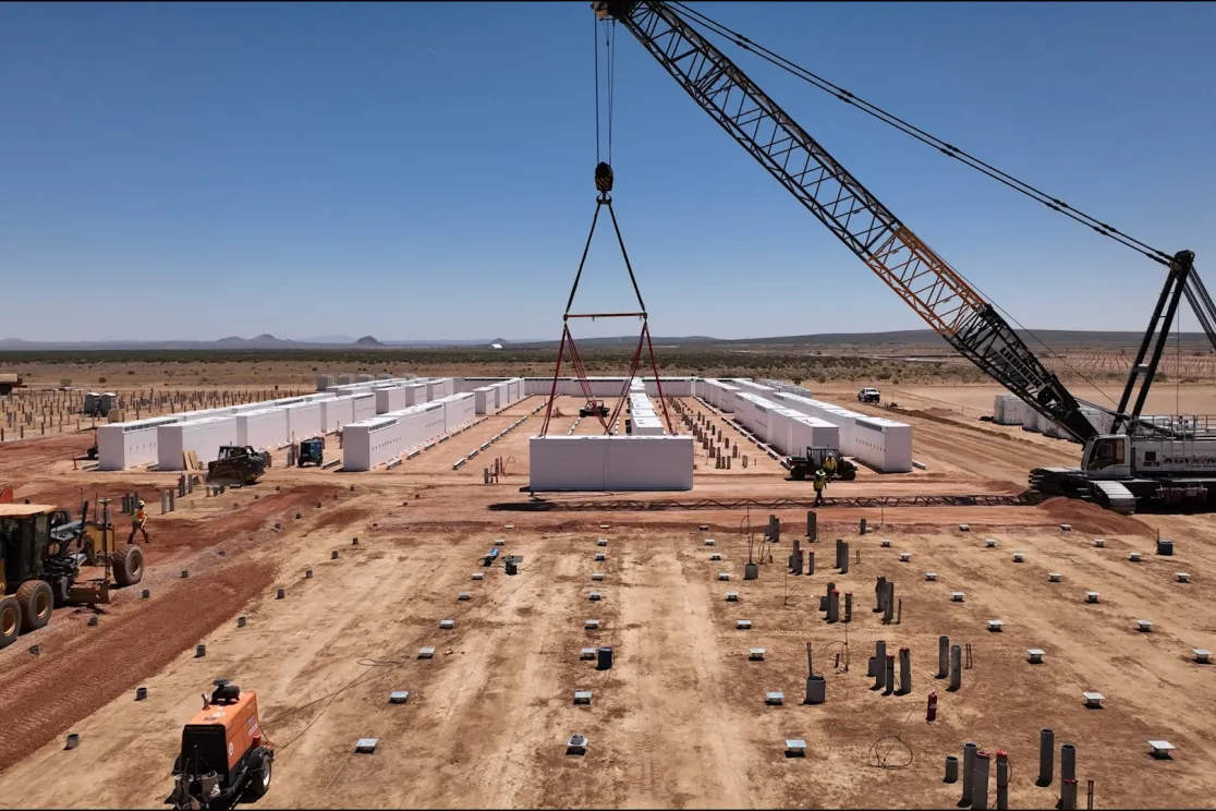 A construction site in a desert landscape with a large crane lifting a white rectangular structure. Rows of similar structures are aligned on the ground. Heavy machinery and workers are visible, with mountains in the distant background under a clear blue sky.