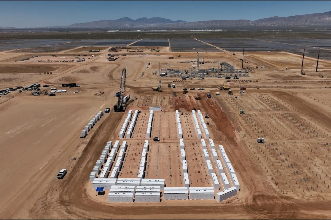 Aerial view of a large construction site in a desert landscape. Rows of white rectangular structures are neatly arranged in the foreground. Construction vehicles and equipment are scattered throughout the site. In the background, there are solar panels and mountains under a clear blue sky.