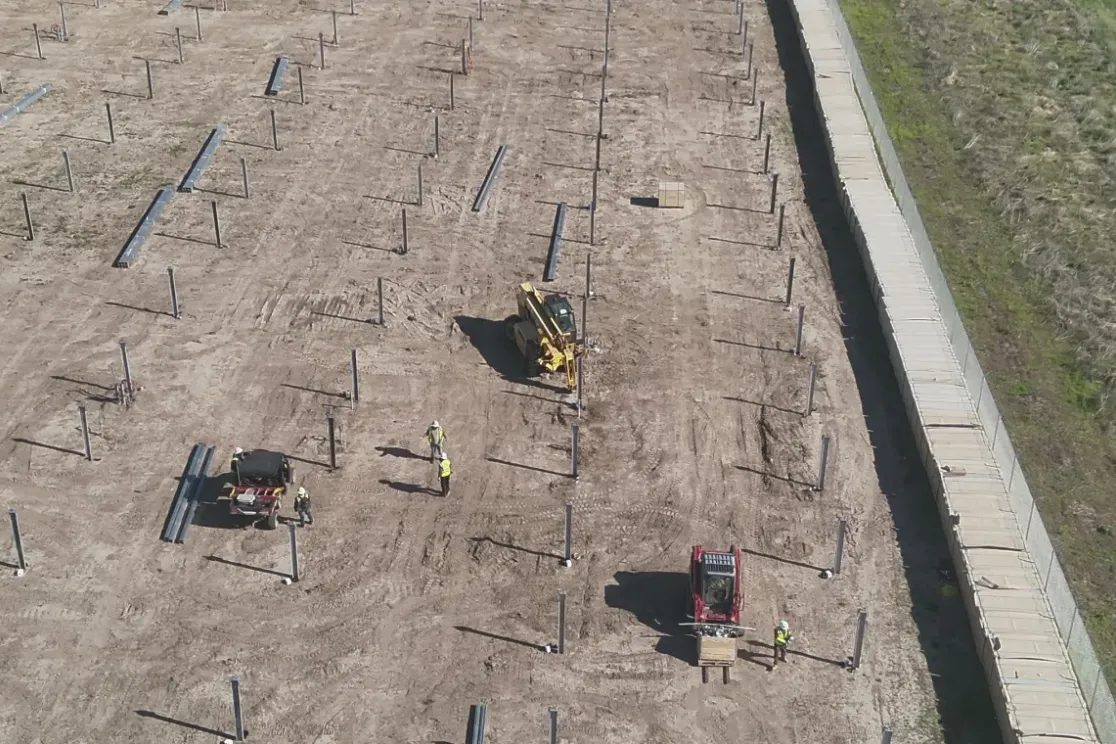 Aerial view of a construction site with rows of metal posts installed in the ground. Workers in high-visibility vests are operating machinery and handling materials. A dirt path runs alongside a green grassy area and a road is visible on the right.