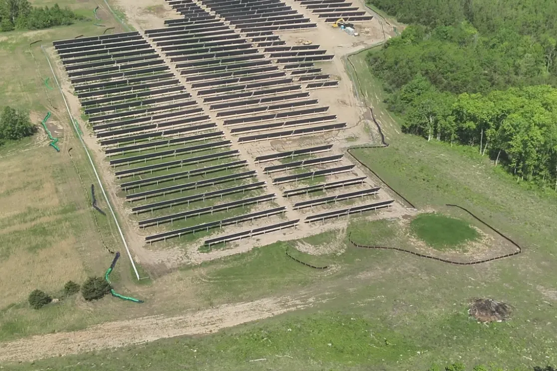 Aerial view of a solar farm with rows of solar panels installed on a cleared piece of land surrounded by dense green forest. The panels are aligned in parallel rows, and there are patches of grass and dirt visible around them.