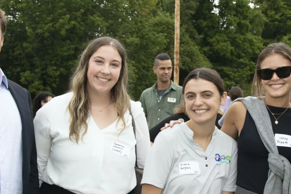 Five people standing outdoors smiling at the camera. They are wearing name tags and casual clothing. Trees and other people are visible in the background.