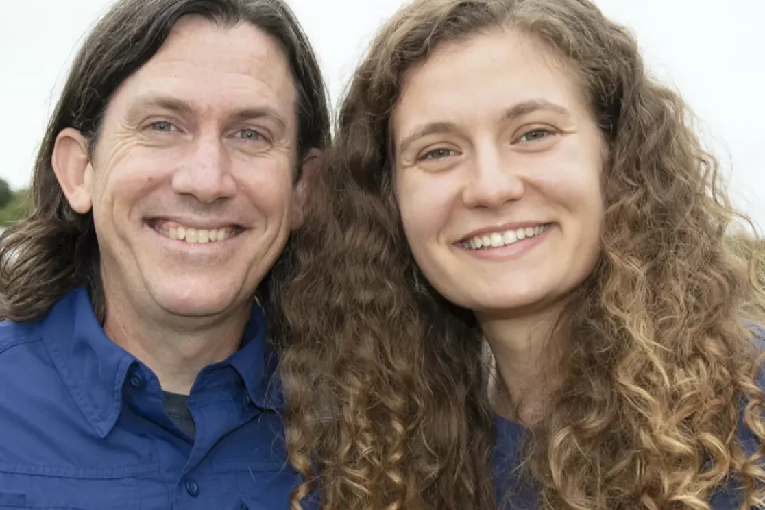 A man and woman smiling outdoors with a blurred background of trees and a building. The man has long hair and is wearing a blue shirt. The woman has curly hair and is also wearing a blue shirt.