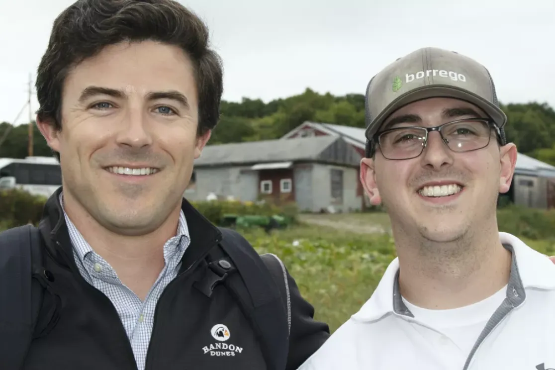 Two men smiling outdoors in front of a barn and trees. One wears a dark jacket with a logo, the other a cap and glasses.