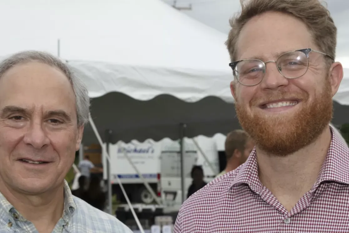 Two men smiling at an outdoor event with a white tent in the background. The man on the left has gray hair and is wearing a plaid shirt, while the man on the right has red hair, a beard, glasses, and is wearing a checkered shirt. Other people and trees are visible in the background.