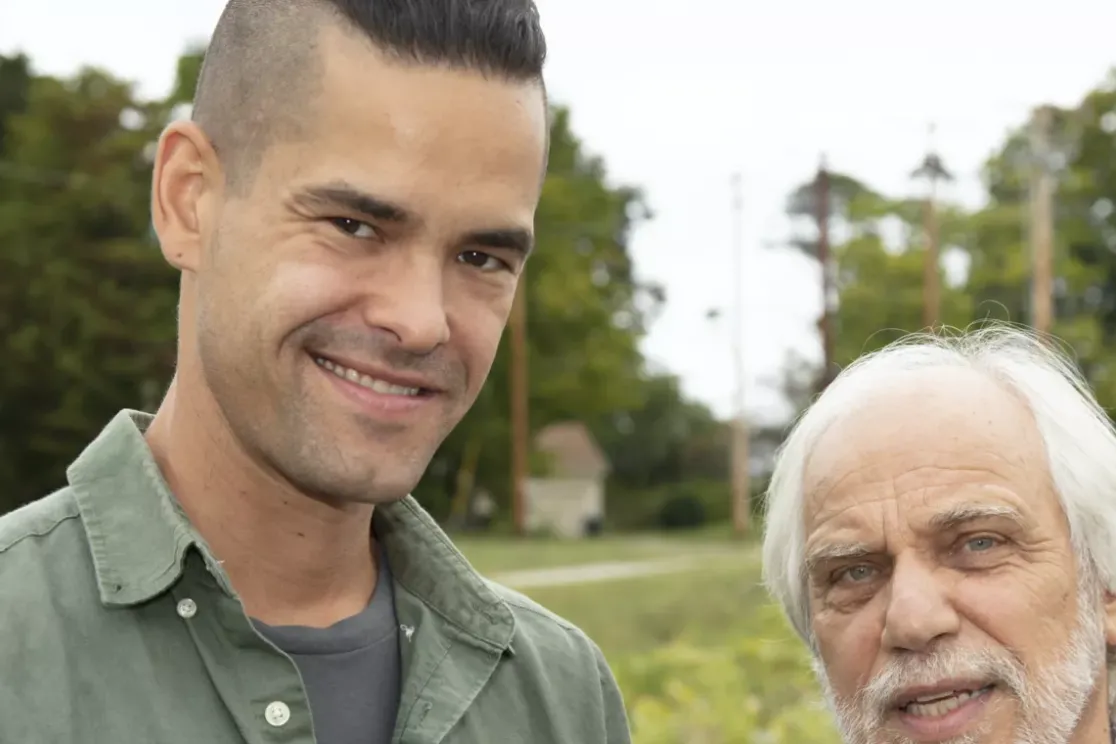 Two men are standing outdoors in a field with trees in the background. The man on the left has short dark hair and is wearing a green shirt. The man on the right has white hair and a beard, wearing a light-colored shirt. Both are smiling softly at the camera.