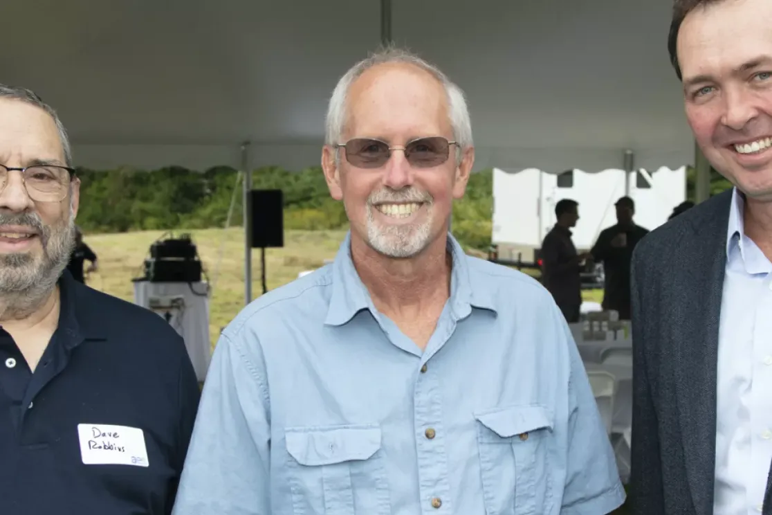 Three men standing under a tent at an outdoor event. The man on the left is wearing a navy polo shirt and glasses. The man in the middle is wearing a light blue shirt and sunglasses. The man on the right is wearing a dark suit jacket over a light blue shirt. They are all smiling, and there are people and tables in the background.