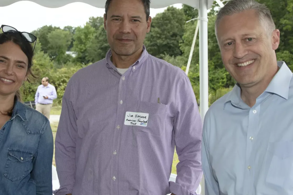 Three people standing under a tent at an outdoor event. The person on the left wears a denim shirt and sunglasses on their head. The middle person wears a purple shirt, and the person on the right wears a light blue shirt. A truck and greenery are visible in the background.