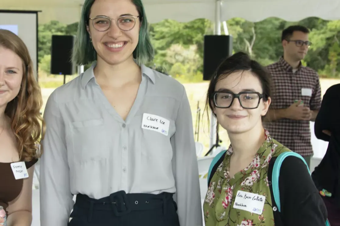 Three people smiling at the camera under a tent during an outdoor event. They are wearing name tags. In the background, a group of people are conversing and a projector screen is visible.