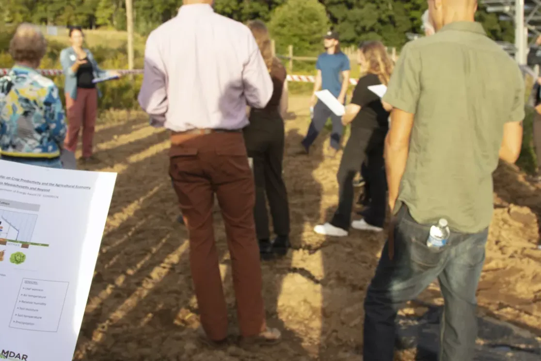 A group of people standing on a dirt path outdoors, holding papers and listening to a speaker. The scene is set near a field with greenery in the background. One person in the foreground holds a paper with a chart and text about impacts on agriculture.
